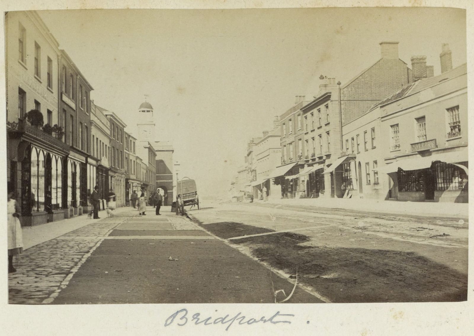 A photograph of a wide town street with shop awnings and a few people.
