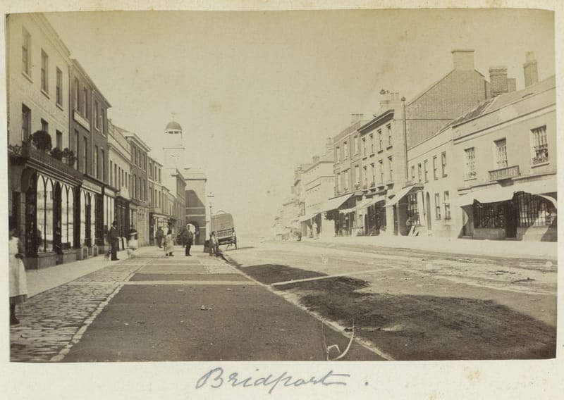 A photograph of a wide town street with shop awnings and a few people.