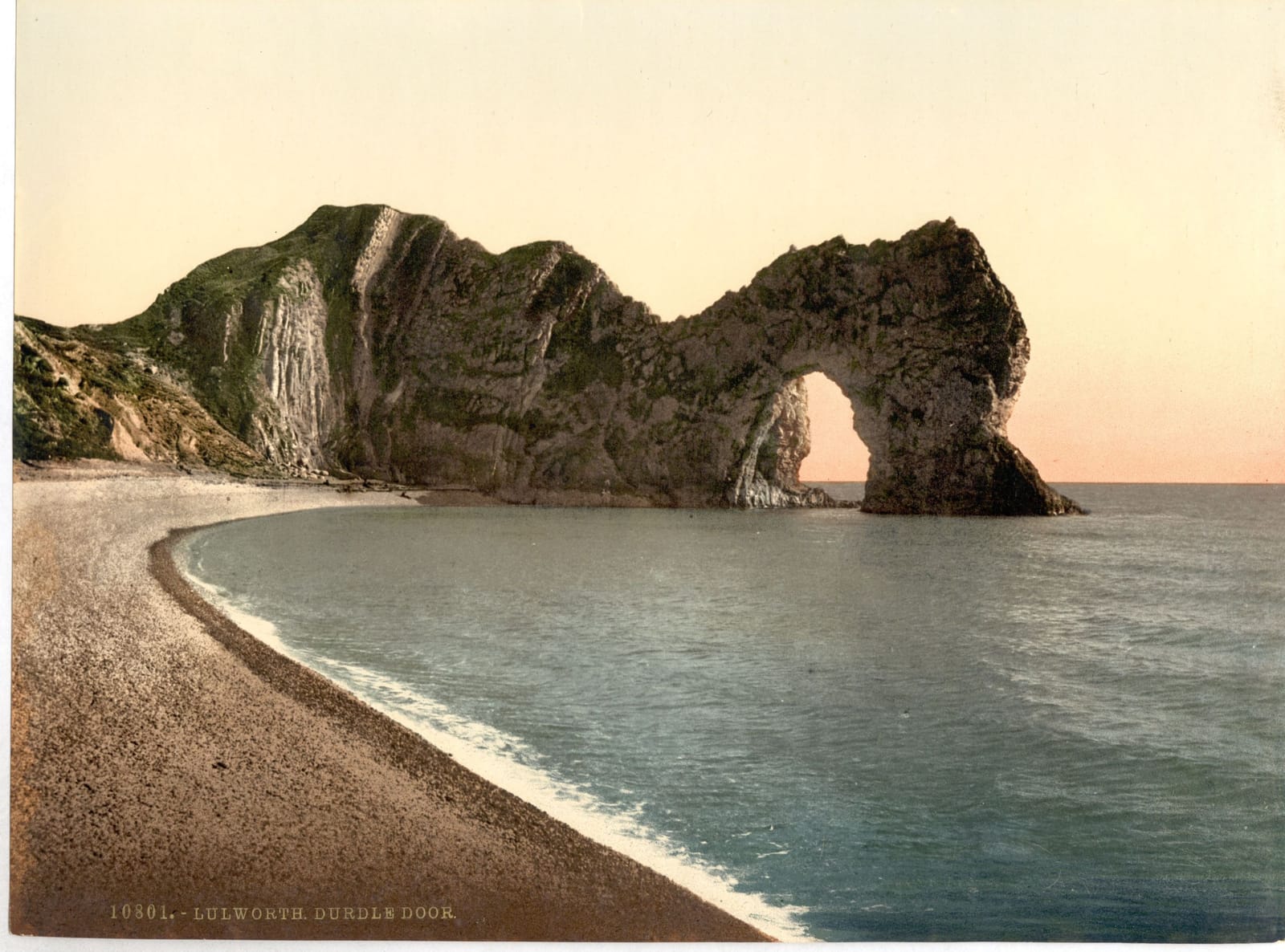 The limestone arch of Durdle Door standing out into a calm sea, with a shingle beach curving towards it.