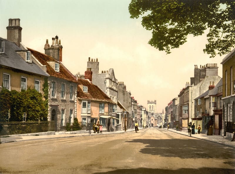 A wide town street of brick and stone houses, with a church tower at the far end.