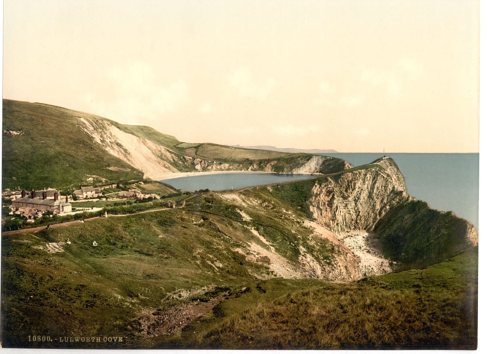 The round bay of Lulworth Cove seen from the green cliff above it, with white chalk and the open sea beyond.