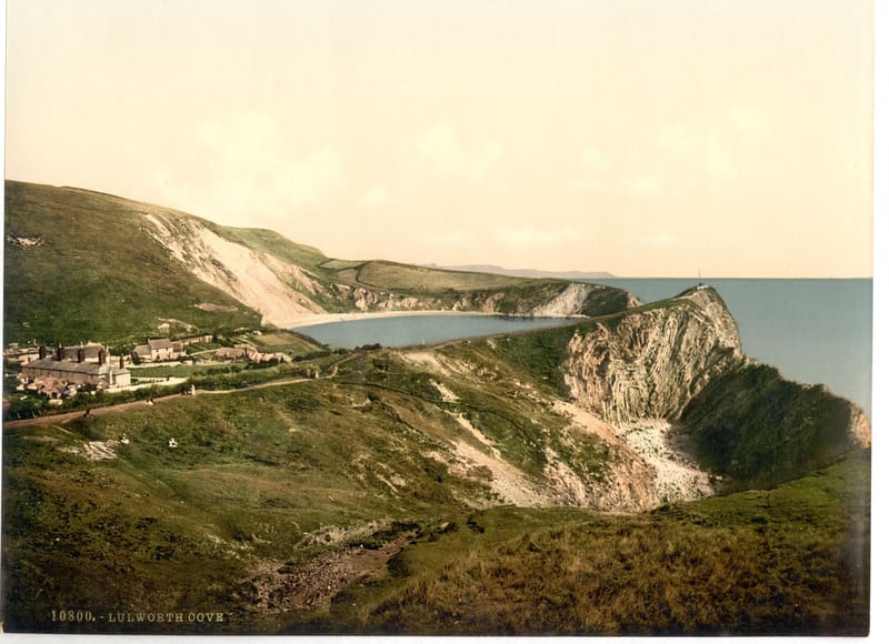 The round bay of Lulworth Cove seen from the green cliff above it, with white chalk and the open sea beyond.