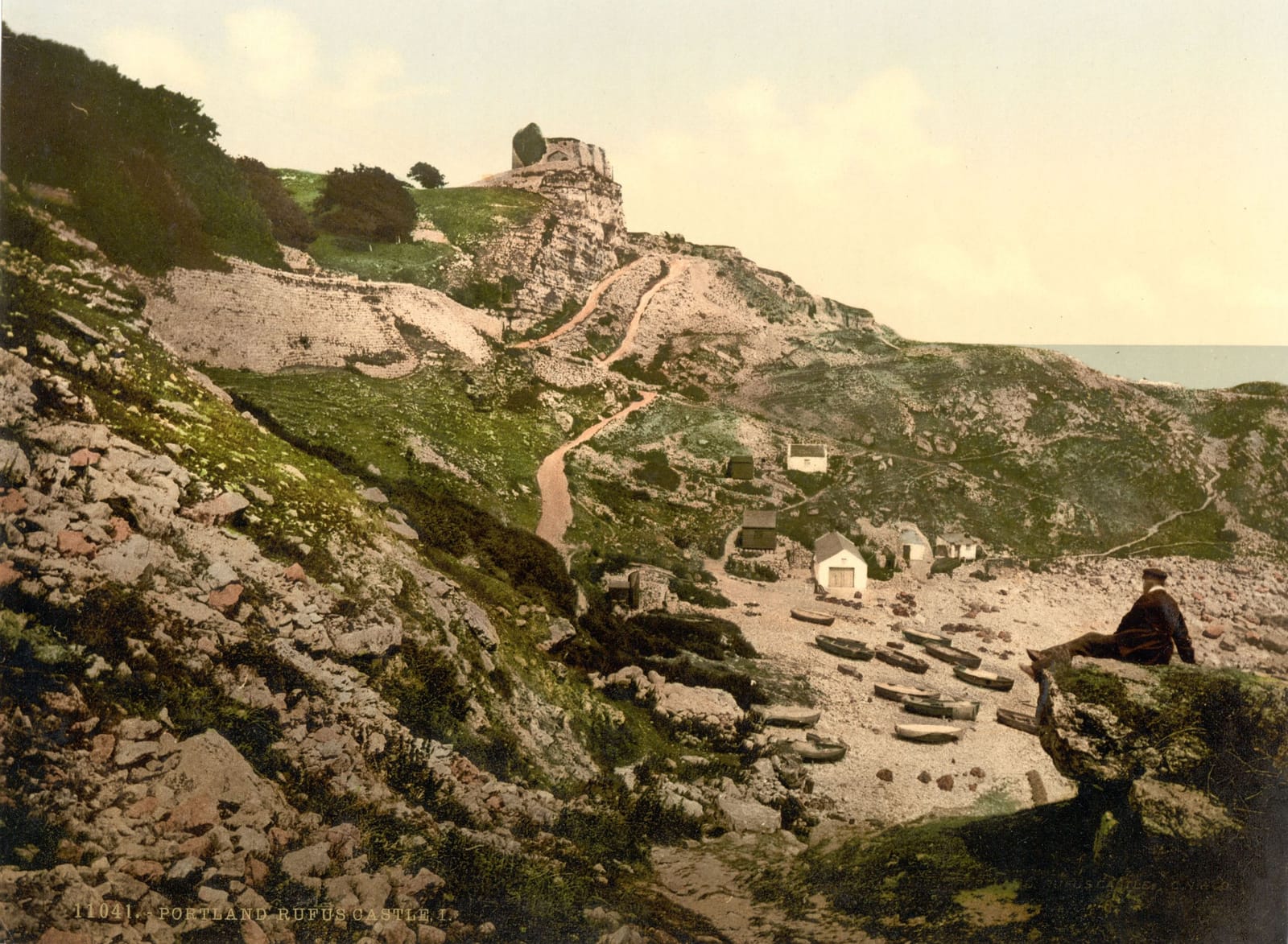 The ruined keep of Rufus Castle perched high on a rocky, overgrown slope above a cove.