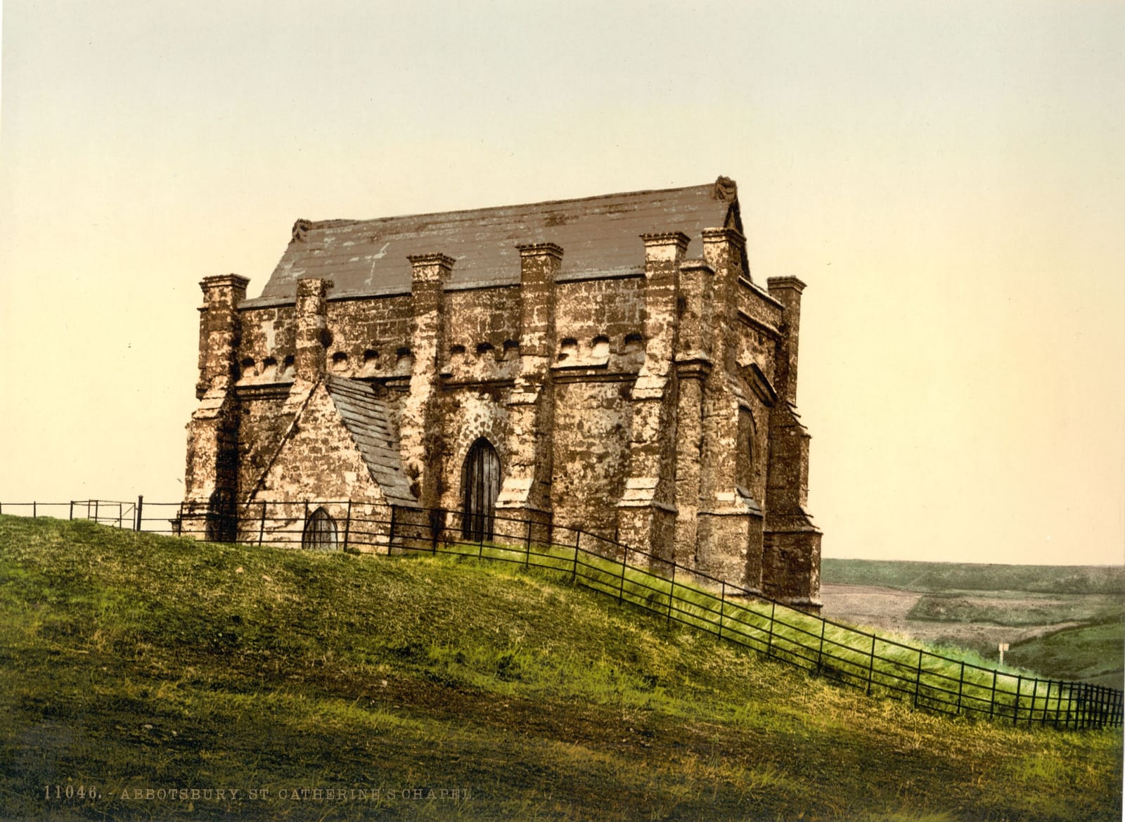 A solid stone chapel standing alone on a grassy hilltop, with fields and the sea in the distance.