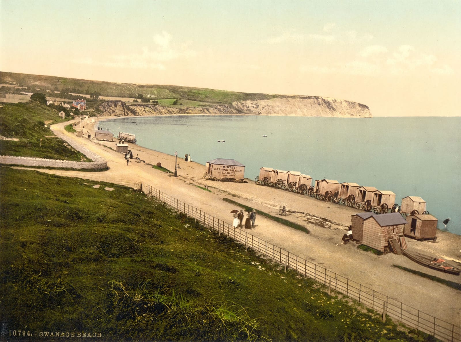 A promenade above a curving beach lined with bathing huts, with the bay and headland beyond.
