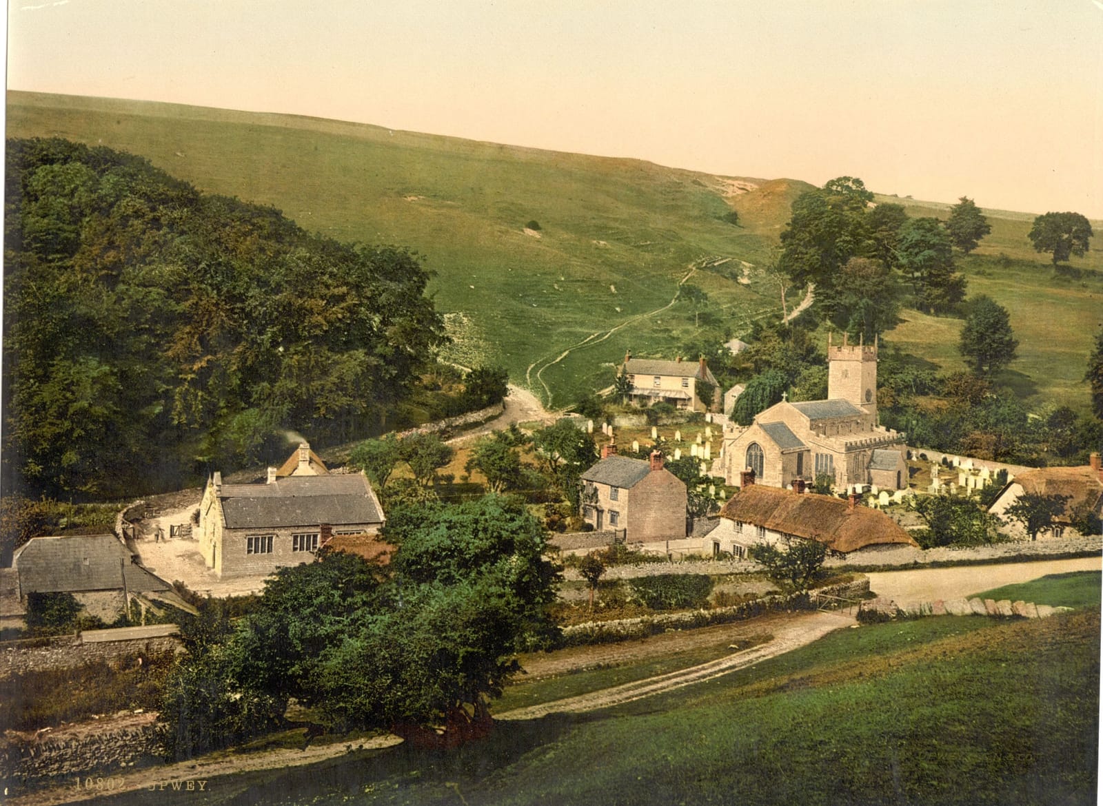 Cottages and trees in a green valley below a steep open down.