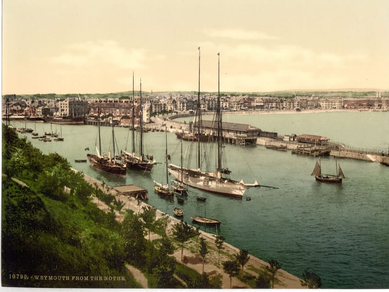 Sailing ships moored in Weymouth harbour, with the town and the long curve of the bay behind.