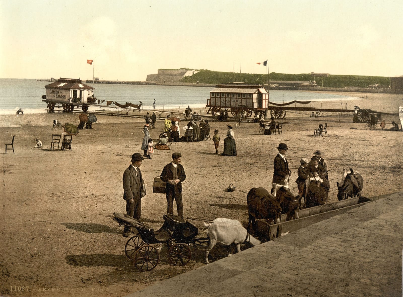 People on Weymouth sands among the bathing machines, with the headland across the bay.